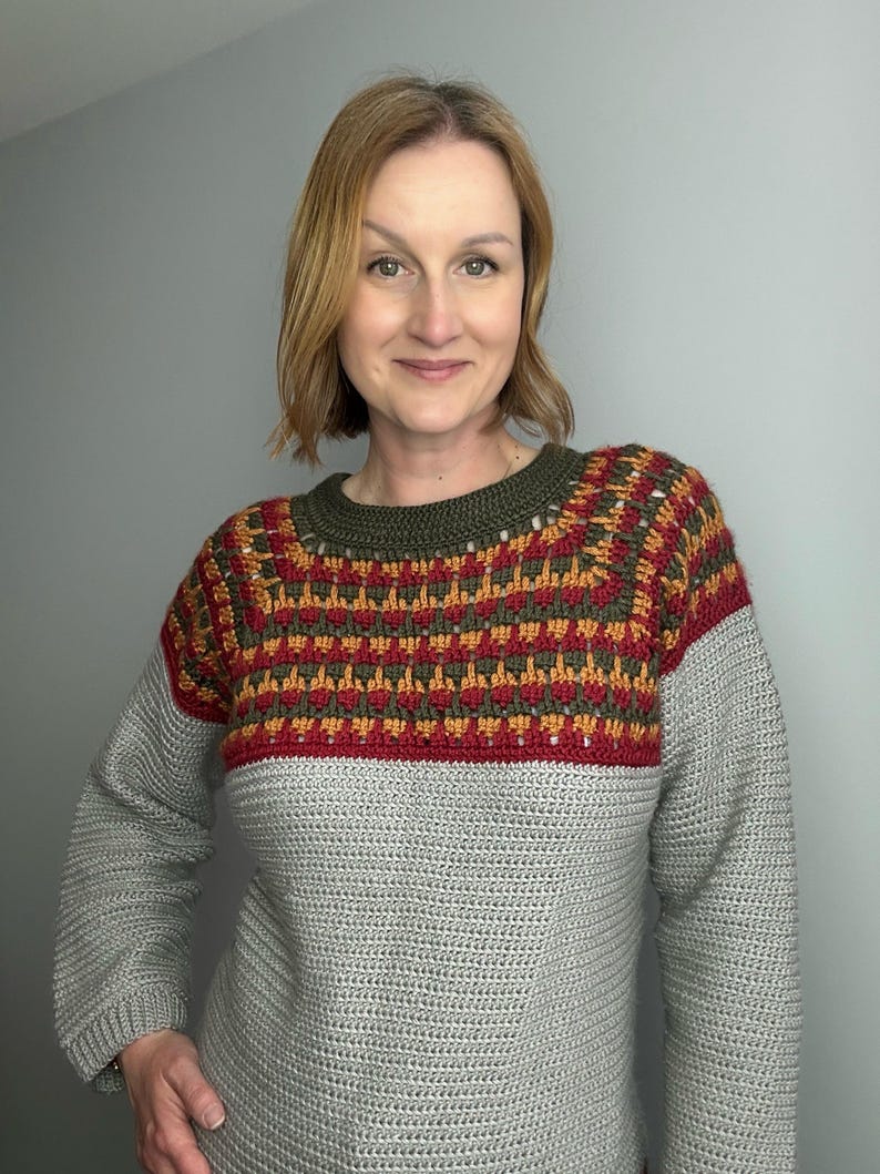 May include: A grey crochet sweater with a colour block design. The upper section features a pattern of red, orange, and green, with a dark green neckline. The sleeves and lower body are a solid grey colour.