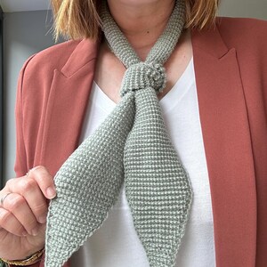 May include: A light green, hand-knitted scarf with a textured pattern. The scarf is tied in a knot at the neck, with the ends hanging down. The scarf is worn over a white shirt and a rust-colored blazer.