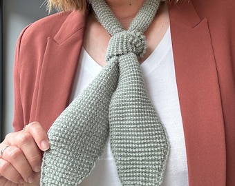May include: A light green, hand-knitted scarf with a textured pattern. The scarf is tied in a knot at the neck, with the ends hanging down. The scarf is worn over a white shirt and a rust-colored blazer.