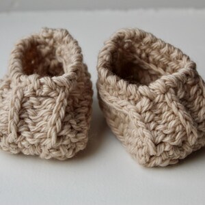 a pair of crochet baby booties in cream color on a plain white background with textured stitches made from a flat rectangle suitable for newborn baby boy or girl for baby shower gift as dyi baby shoes