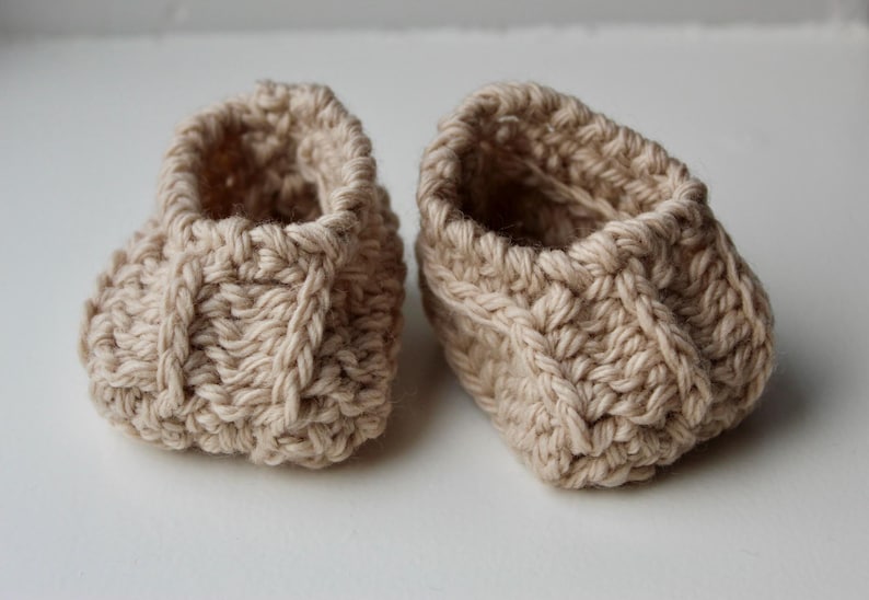 a pair of crochet baby booties in cream color on a plain white background with textured stitches made from a flat rectangle suitable for newborn baby boy or girl for baby shower gift as dyi baby shoes