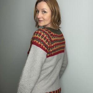 May include: A grey crochet sweater with a colour block design. The upper section features a pattern of red, orange, and green, while the sleeves and lower body are solid grey. The sweater has a crew neck and long sleeves.