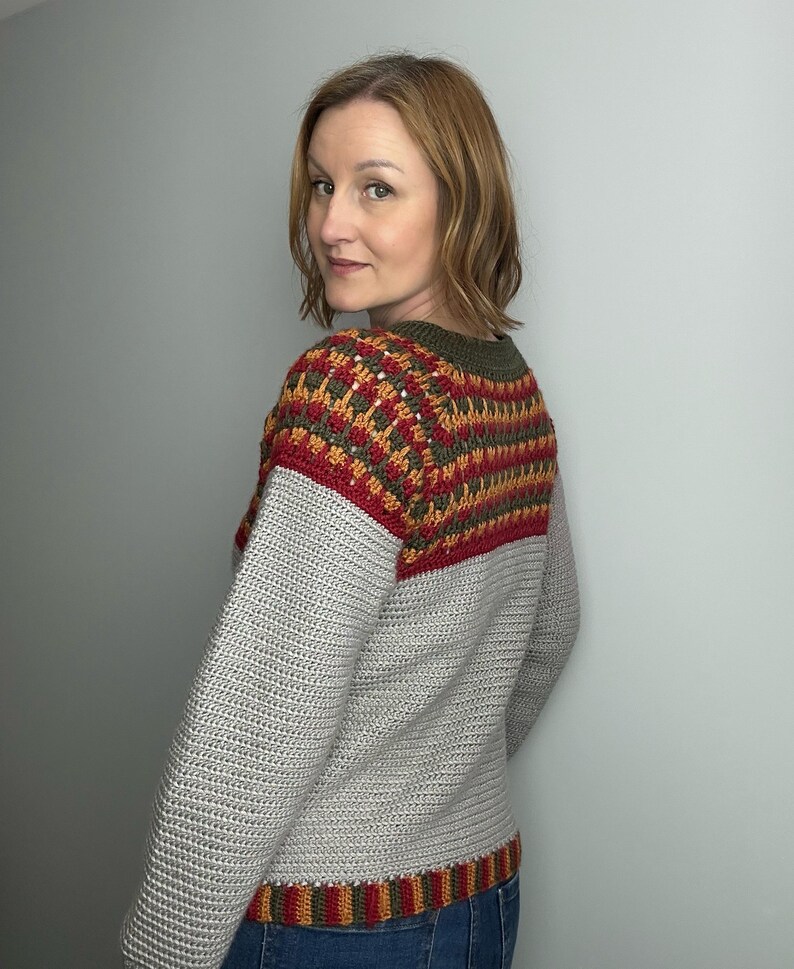 May include: A grey crochet sweater with a colour block design. The upper section features a pattern of red, orange, and green, while the sleeves and lower body are solid grey. The sweater has a crew neck and long sleeves.
