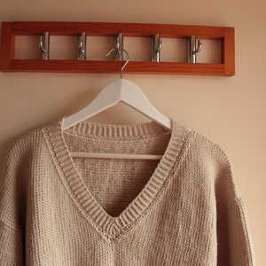 A beige, hand-crocheted V-neck sweater hanging on a white hanger, which is hooked onto a wooden wall-mounted coat rack with five metal hooks. The background wall is painted in a light cream color.