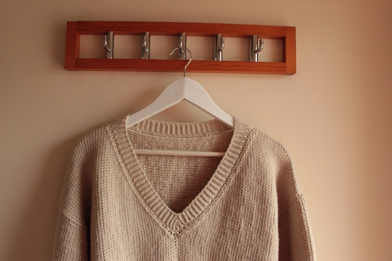 A beige, hand-crocheted V-neck sweater hanging on a white hanger, which is hooked onto a wooden wall-mounted coat rack with five metal hooks. The background wall is painted in a light cream color.