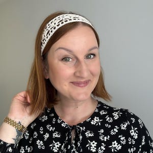 May include: A cream-colored, crocheted headband. The headband is worn on the head. The person in the image is wearing a black top with a white floral pattern. The person has short, reddish-brown hair.