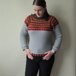May include: A grey crochet sweater with a colour block design. The upper chest and sleeves feature a pattern of red, orange, and green. The sweater has long sleeves and a crew neck. The bottom hem also has a striped pattern.