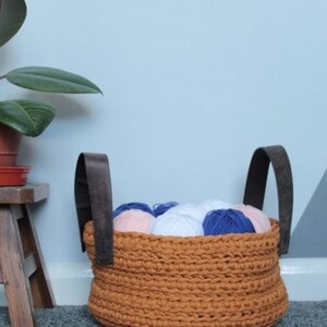 May include: A brown crocheted storage basket with leather handles filled with balls of yarn in pink, white, and blue.