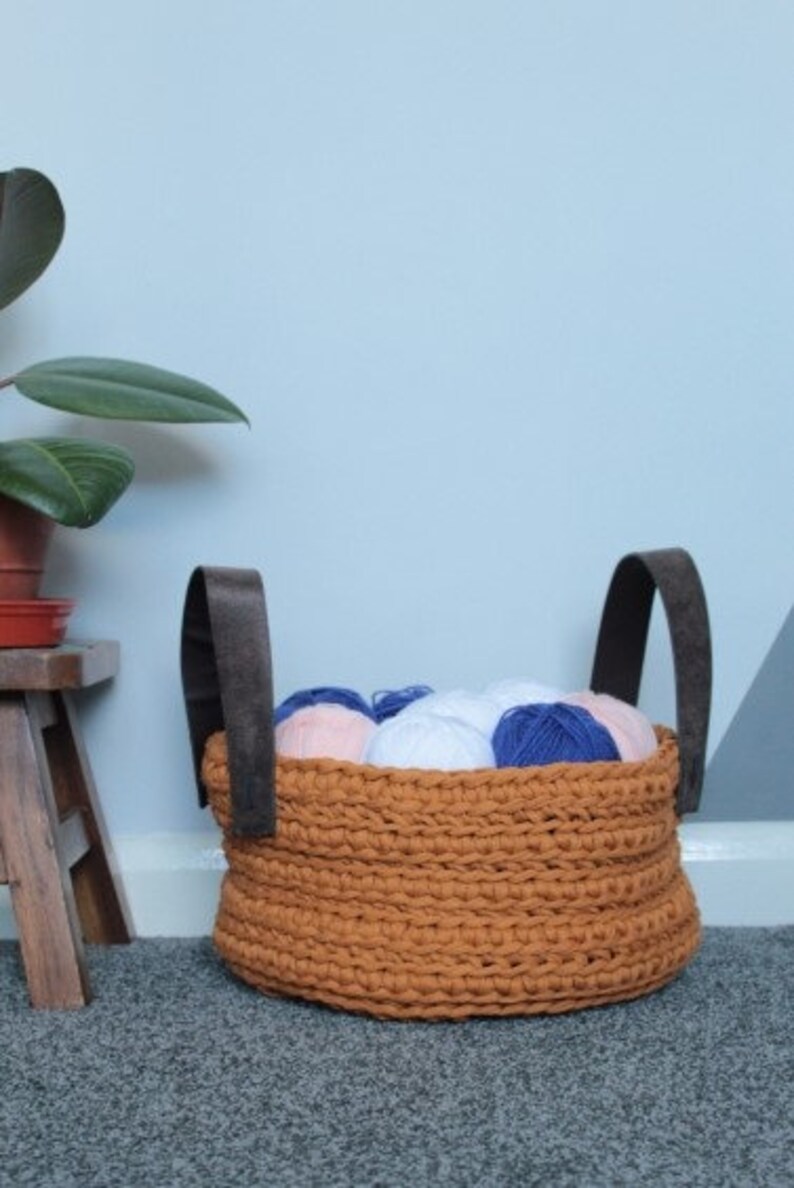 May include: A brown crocheted storage basket with leather handles filled with balls of yarn in pink, white, and blue.