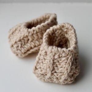 a pair of crochet baby booties in cream color on a plain white background with textured stitches made from a flat rectangle suitable for newborn baby boy or girl for baby shower gift as dyi baby shoes