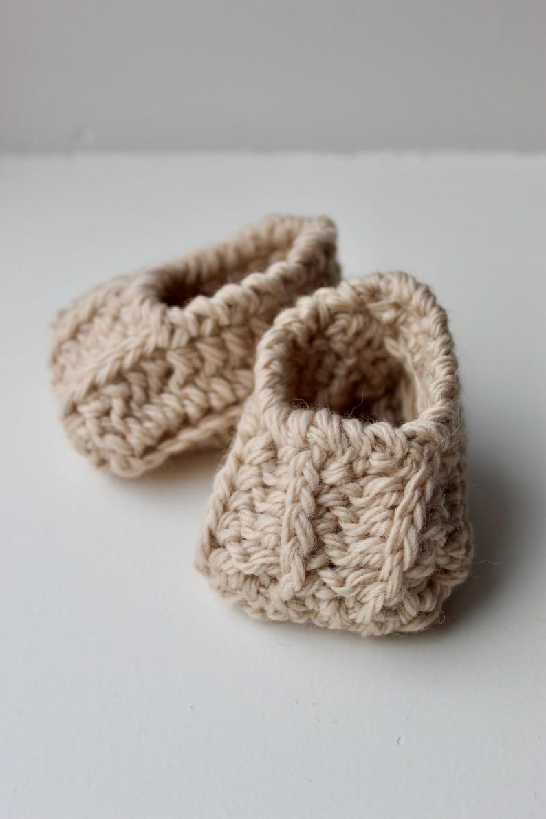 a pair of crochet baby booties in cream color on a plain white background with textured stitches made from a flat rectangle suitable for newborn baby boy or girl for baby shower gift as dyi baby shoes