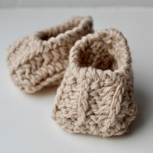 a pair of crochet baby booties in cream color on a plain white background with textured stitches made from a flat rectangle suitable for newborn baby boy or girl for baby shower gift as dyi baby shoes