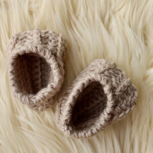 a pair of crochet baby booties in cream color on a plain white background with textured stitches made from a flat rectangle suitable for newborn baby boy or girl for baby shower gift as dyi baby shoes