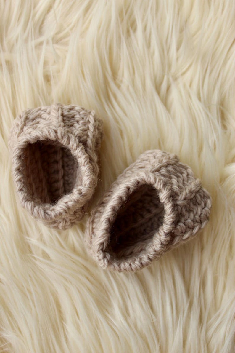 a pair of crochet baby booties in cream color on a plain white background with textured stitches made from a flat rectangle suitable for newborn baby boy or girl for baby shower gift as dyi baby shoes