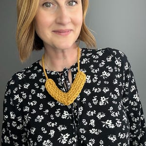 Op de afbeelding: Mosterdgele gehaakte ketting in V-vorm. De ketting is gemaakt van dik garen en heeft een textuur. De vrouw draagt een zwarte blouse met bloemenprint.
