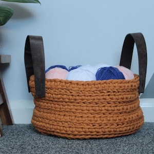 May include: A brown crocheted basket with leather handles filled with balls of yarn in pink, white, and blue.
