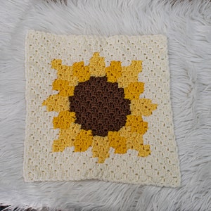May include: A crocheted square pillow with a sunflower design. The sunflower is made with yellow, brown, and white yarn.