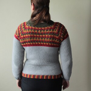 May include: A grey crochet sweater with long sleeves. The upper section has a colourful pattern in red, orange, and yellow. The neckline and hem have a striped design. A handmade, cosy garment.