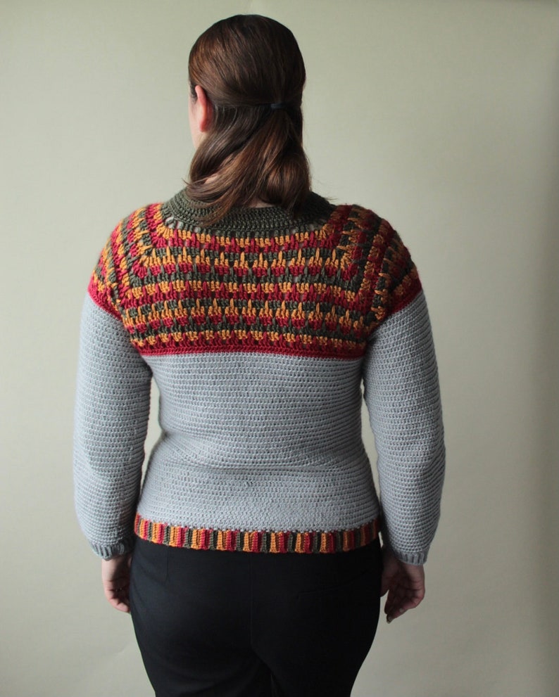 May include: A grey crochet sweater with long sleeves. The upper section has a colourful pattern in red, orange, and yellow. The neckline and hem have a striped design. A handmade, cosy garment.