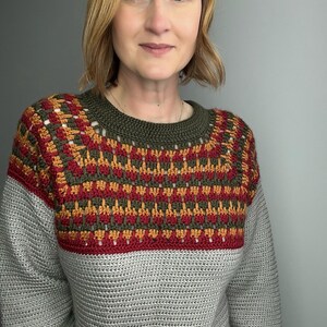 May include: A grey crochet sweater with a patterned yoke in red, gold, and olive green. The yoke features a geometric design. The sweater has long sleeves and a crew neck. The model is wearing the sweater.