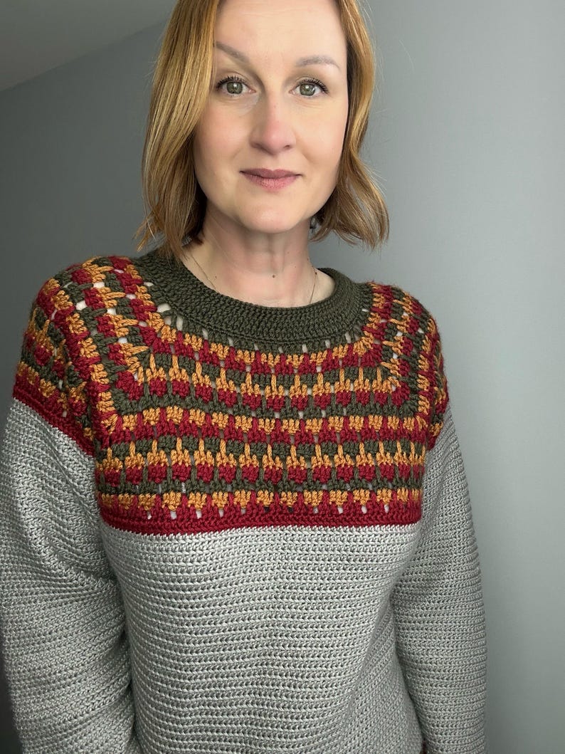 May include: A grey crochet sweater with a patterned yoke in red, gold, and olive green. The yoke features a geometric design. The sweater has long sleeves and a crew neck. The model is wearing the sweater.