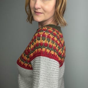 May include: A grey crochet jumper with a colour block design on the shoulders. The colour block features red, gold, and olive green geometric patterns. The jumper has a crew neck and long sleeves.