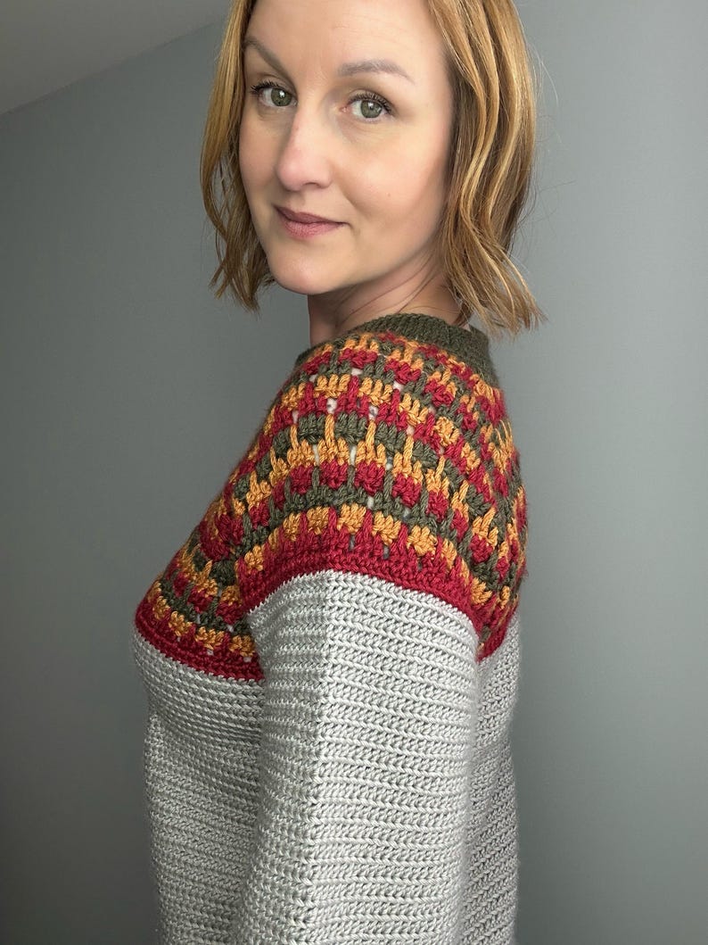 May include: A grey crochet jumper with a colour block design on the shoulders. The colour block features red, gold, and olive green geometric patterns. The jumper has a crew neck and long sleeves.