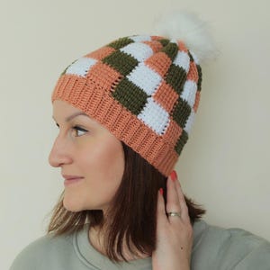 May include: A crocheted beanie featuring a checkered pattern in alternating squares of terracotta orange, olive green, and white. The hat has a solid terracotta orange ribbed brim and is topped with a fluffy white faux fur pom-pom.