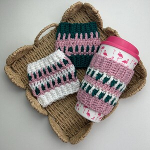May include: Three handmade crochet cup sleeves in a woven basket. One sleeve is on a travel mug. The sleeves are pink, white, and green with a geometric pattern. The travel mug has a pink lid and a flamingo design.