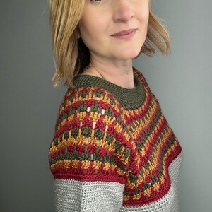May include: A handmade crochet sweater with a colour block design. The top section features a pattern of red, gold, and olive green, while the lower section is solid grey. The sweater has a crew neck and short sleeves.
