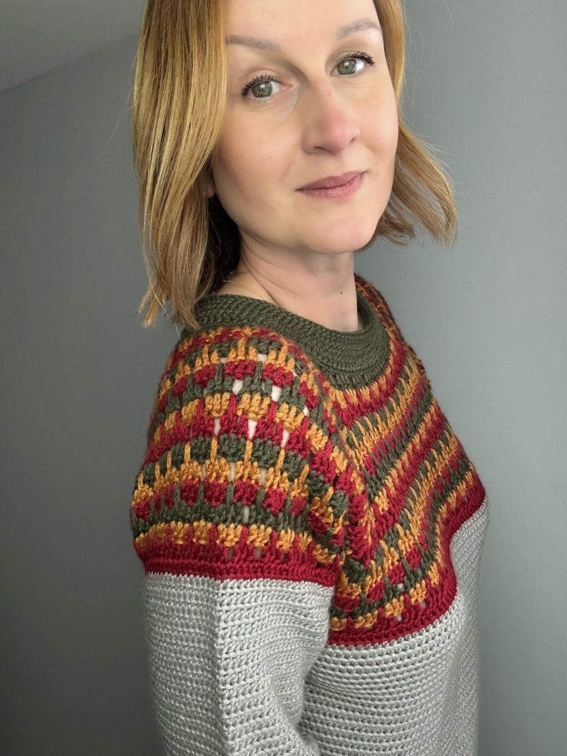 May include: A handmade crochet sweater with a colour block design. The top section features a pattern of red, gold, and olive green, while the lower section is solid grey. The sweater has a crew neck and short sleeves.