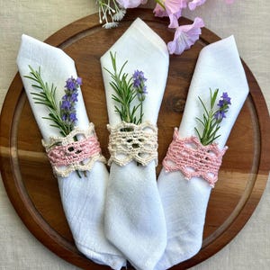 May include: Three white cloth napkins, each adorned with a sprig of lavender and rosemary, are held by crochet napkin rings. Two rings are pink, and one is cream-colored. The napkins rest on a wooden serving board.