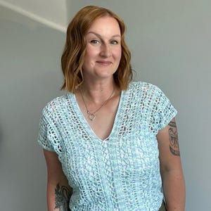 Puede incluir: Una blusa de crochet azul claro con cuello en V y mangas cortas. La blusa se combina con pantalones verde oliva. El diseño de crochet es abierto y aireado. La mujer lleva un collar.