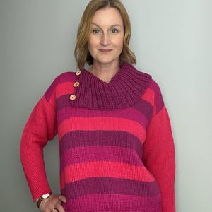 Könnte beinhalten: Ein gestreifter Pullover in Rosa- und Lilatönen mit einem Rollkragen und drei Holzknöpfen. Der Pullover hat lange Ärmel und ist aus einem dicken Strickmaterial gefertigt. Das Farbschema umfasst Rosa- und Lilatöne.