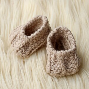 a pair of crochet baby booties in cream color on a plain white background with textured stitches made from a flat rectangle suitable for newborn baby boy or girl for baby shower gift as dyi baby shoes