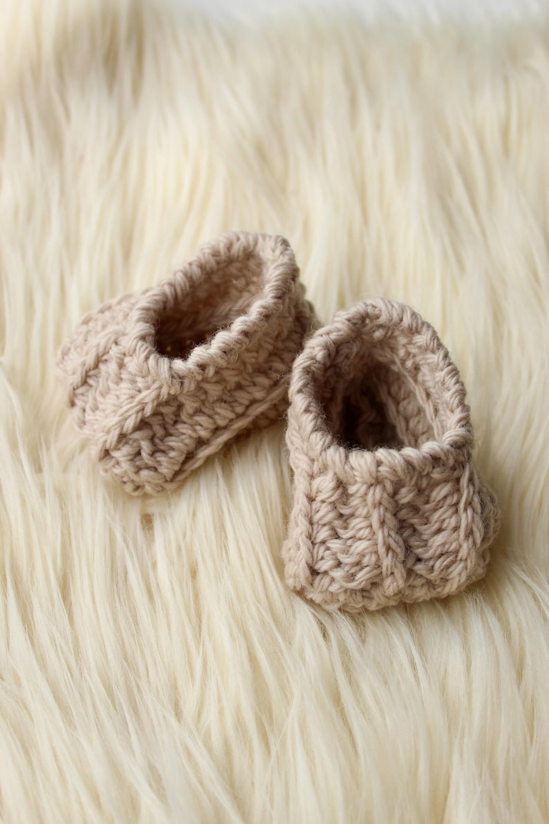 a pair of crochet baby booties in cream color on a plain white background with textured stitches made from a flat rectangle suitable for newborn baby boy or girl for baby shower gift as dyi baby shoes