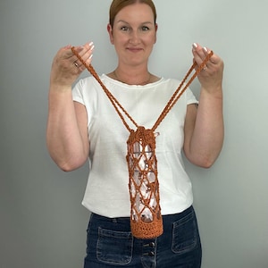 May include: A crocheted water bottle holder in a warm brown hue, featuring a diamond-patterned net design. The holder has a long strap for carrying and is designed to fit a standard-sized water bottle. The overall aesthetic is handmade and rustic.