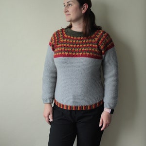 May include: A grey crochet jumper with long sleeves and a crew neck. The yoke features a geometric pattern in red, yellow, and brown. The bottom hem also has a decorative band of the same colours. The jumper is handmade.