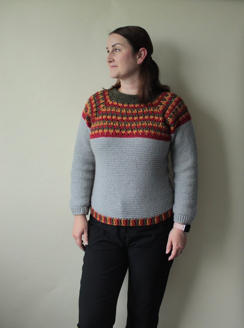 May include: A grey crochet jumper with long sleeves and a crew neck. The yoke features a geometric pattern in red, yellow, and brown. The bottom hem also has a decorative band of the same colours. The jumper is handmade.