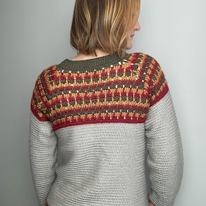 May include: A hand-crocheted sweater in grey, red, yellow, and green. The sweater has a detailed pattern across the upper chest and shoulders, with a solid grey body and sleeves. The neckline and hem are trimmed with a striped pattern.