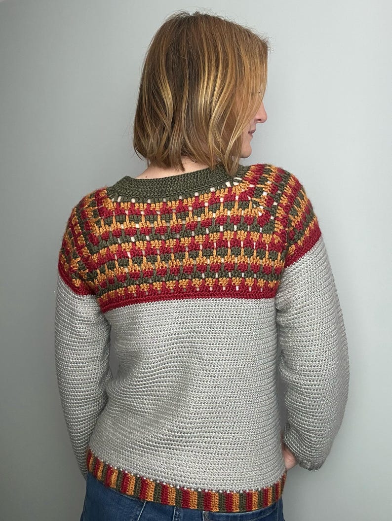 May include: A hand-crocheted sweater in grey, red, yellow, and green. The sweater has a detailed pattern across the upper chest and shoulders, with a solid grey body and sleeves. The neckline and hem are trimmed with a striped pattern.