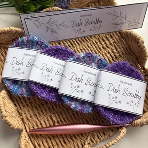 Op de afbeelding: Vier ronde, getextureerde afwassponsjes in paarse en blauwe tinten, elk omwikkeld met een witte papieren band met het label "handmade Dish Scrubby" en florale lijntekeningen. Een metallic roségouden haaknaald ligt onder de sponsjes in een mand.