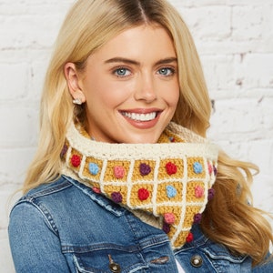 May include: A woman wearing a denim jacket with a crocheted neck warmer featuring a colourful waffle pattern. The neck warmer is made with a variety of colours, including yellow, red, blue, and purple.