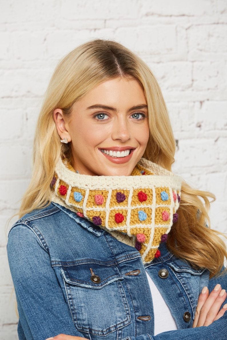 May include: A woman wearing a denim jacket with a crocheted neck warmer featuring a colourful waffle pattern. The neck warmer is made with a variety of colours, including yellow, red, blue, and purple.