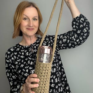 May include: A beige crochet water bottle holder with a long strap. The holder contains a clear glass water bottle with a rose gold lid. The holder is made of a woven, open-stitch design.