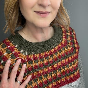May include: A close-up of a crocheted top with a geometric pattern in red, orange, and green. The top has a grey base and a green neckline. The person wearing the top is smiling.