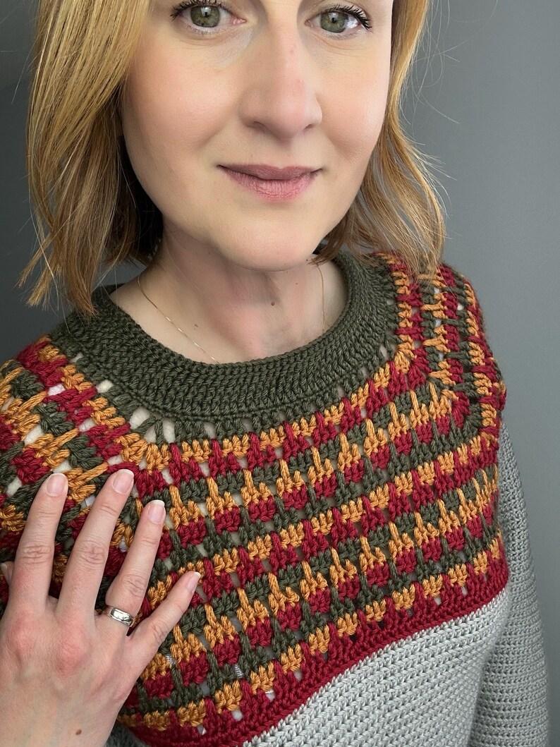 May include: A close-up of a crocheted top with a geometric pattern in red, orange, and green. The top has a grey base and a green neckline. The person wearing the top is smiling.