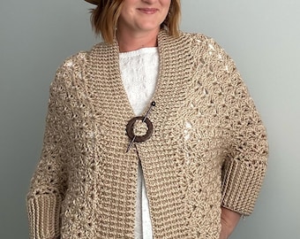 May include: A beige crochet cardigan with a shawl collar and three-quarter sleeves. The cardigan is fastened with a wooden brooch. The person in the image is wearing a brown hat and a white top.