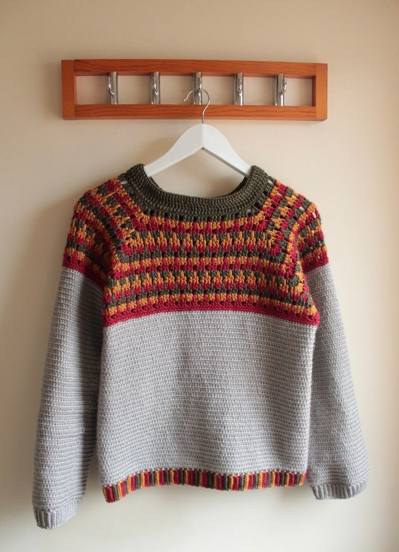May include: A handmade crochet sweater in shades of grey, red, yellow, and olive green hangs on a white hanger. The sweater features a patterned yoke and cuffs, with a solid grey body and sleeves. A wooden coat rack is in the background.
