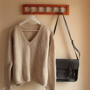 A beige, hand-crocheted V-neck sweater hanging on a white hanger, which is hooked onto a wooden wall-mounted coat rack with five metal hooks. The background wall is painted in a light cream color.